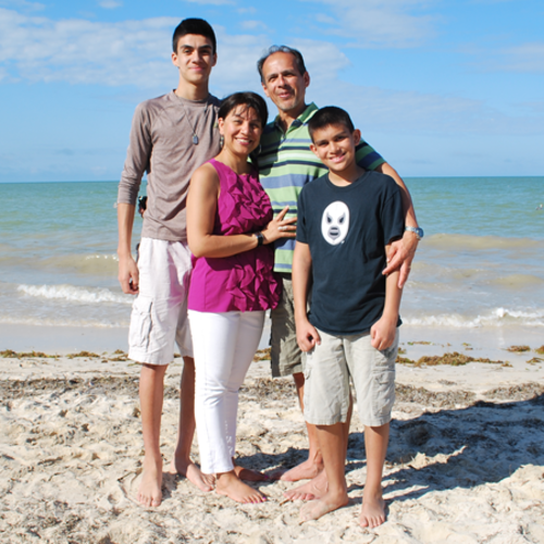 A family of four poses together on a beach with the sea and blue sky in the background.